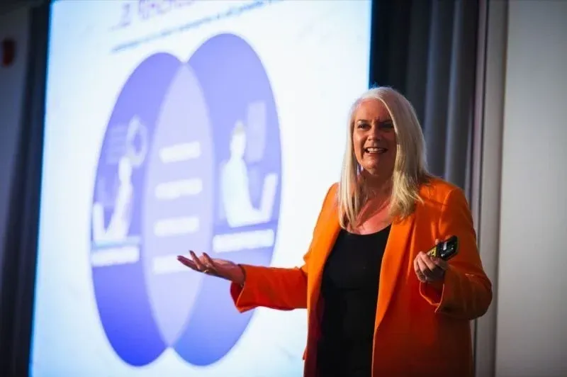 Dr Alison Edgar MBE in an orange blazer, presenting in front of a projected slide showing a Venn diagram.
