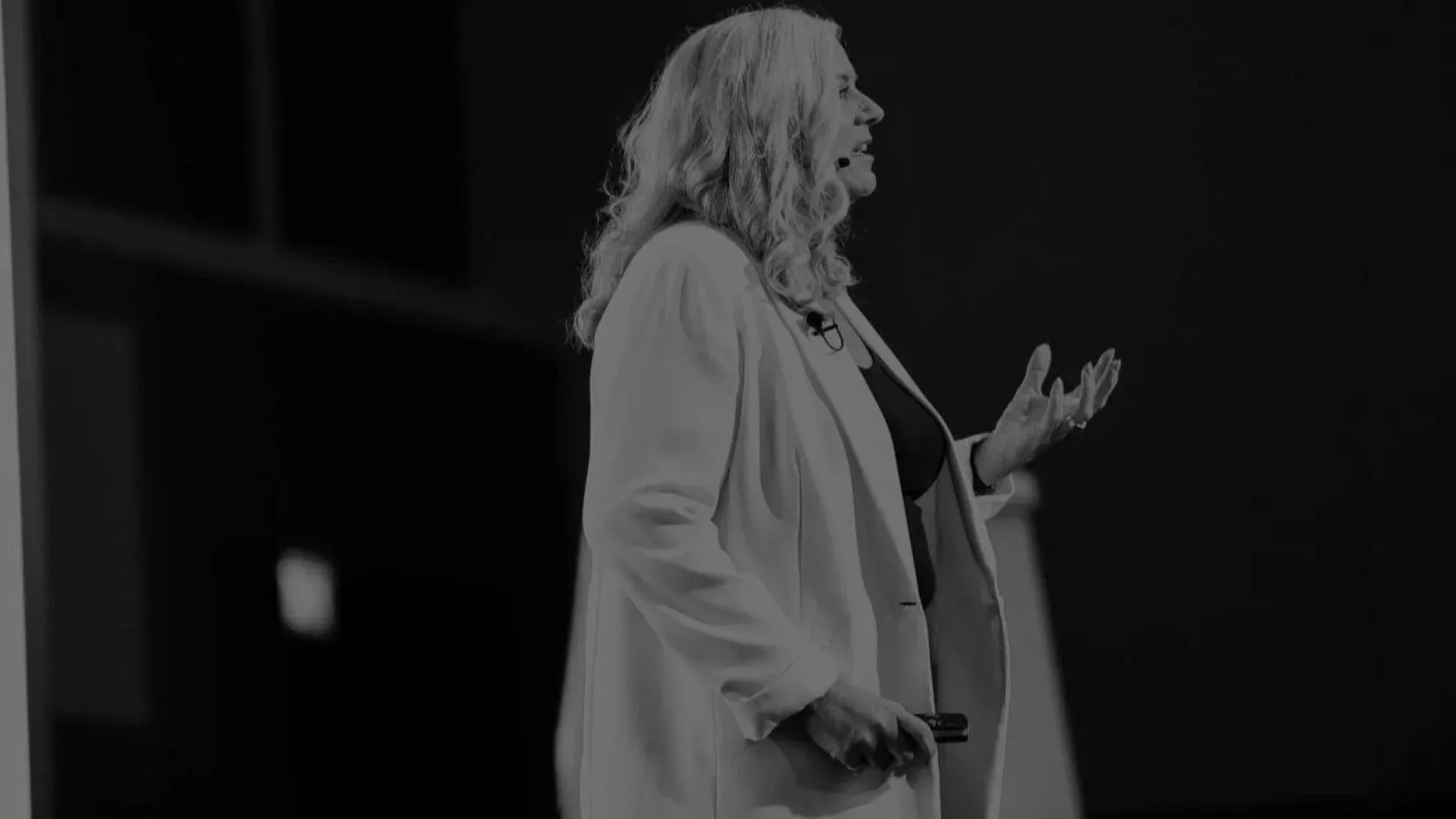 Dr Alison Edgar MBE in profile on a dark stage, mid-sentence with one hand open, in black and white.