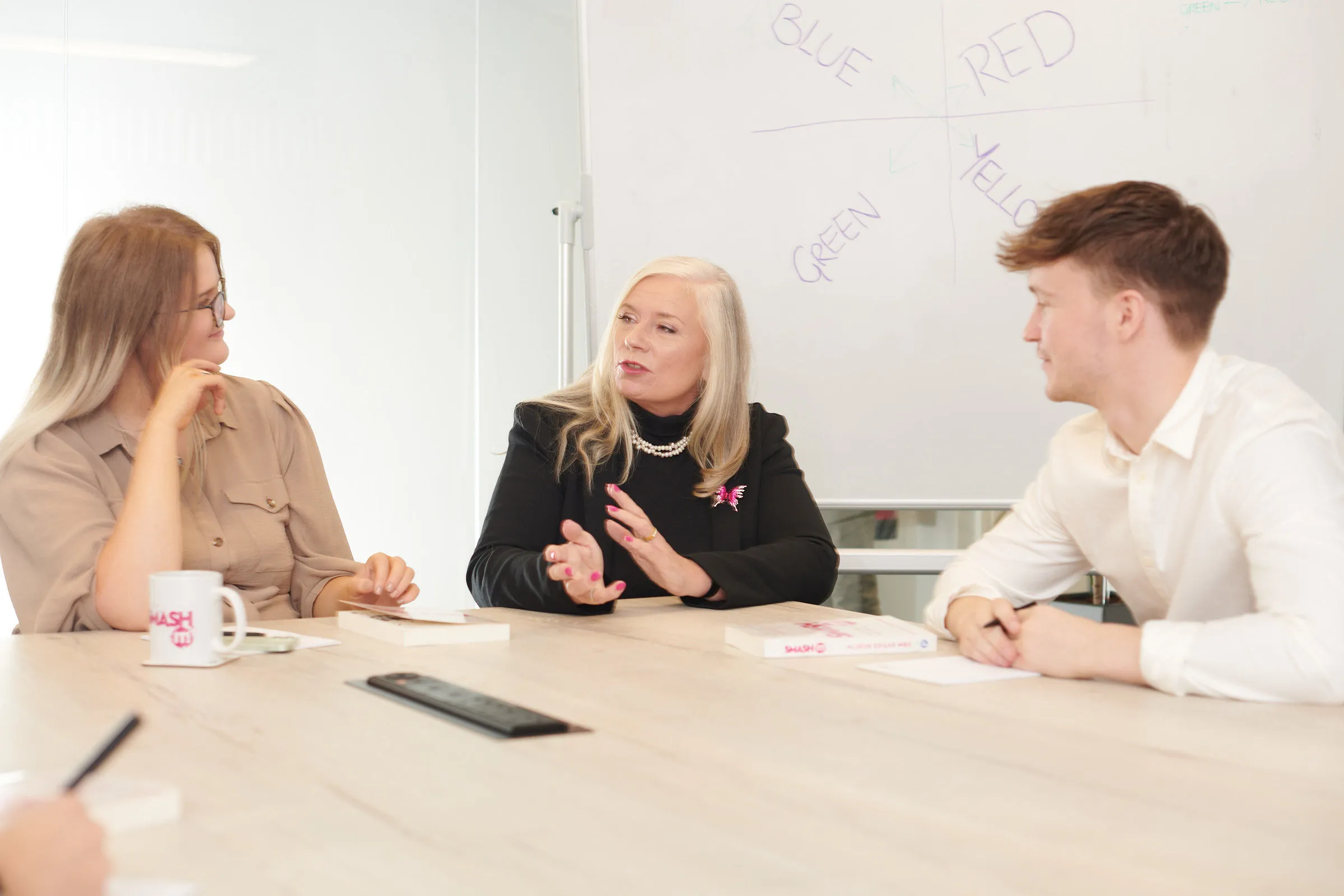 Dr Alison Edgar MBE explaining a behavioural model to two colleagues at a table, colour groupings sketched on the whiteboard behind her.