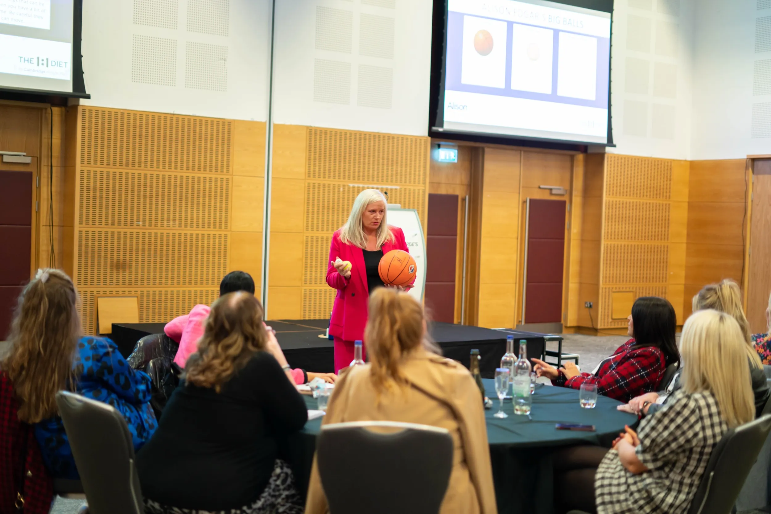 Dr Alison Edgar MBE in a pink suit on stage holding a basketball and a tennis ball, her Big Balls® slide on the screen behind her, delegates watching from round tables.