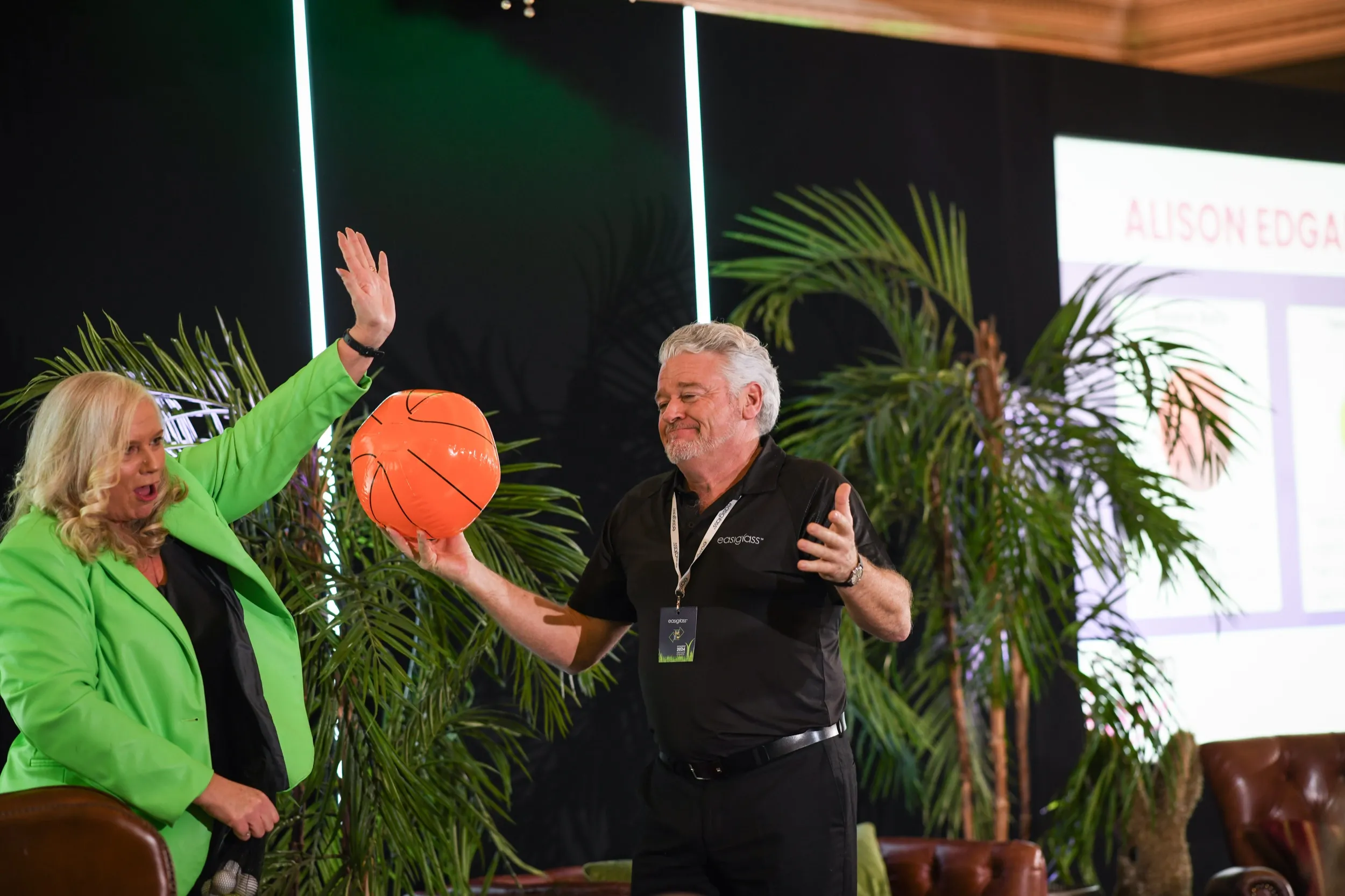 Dr Alison Edgar MBE in a green suit, arm raised, on stage at an EasiGrass event as a delegate holds up an orange ball.
