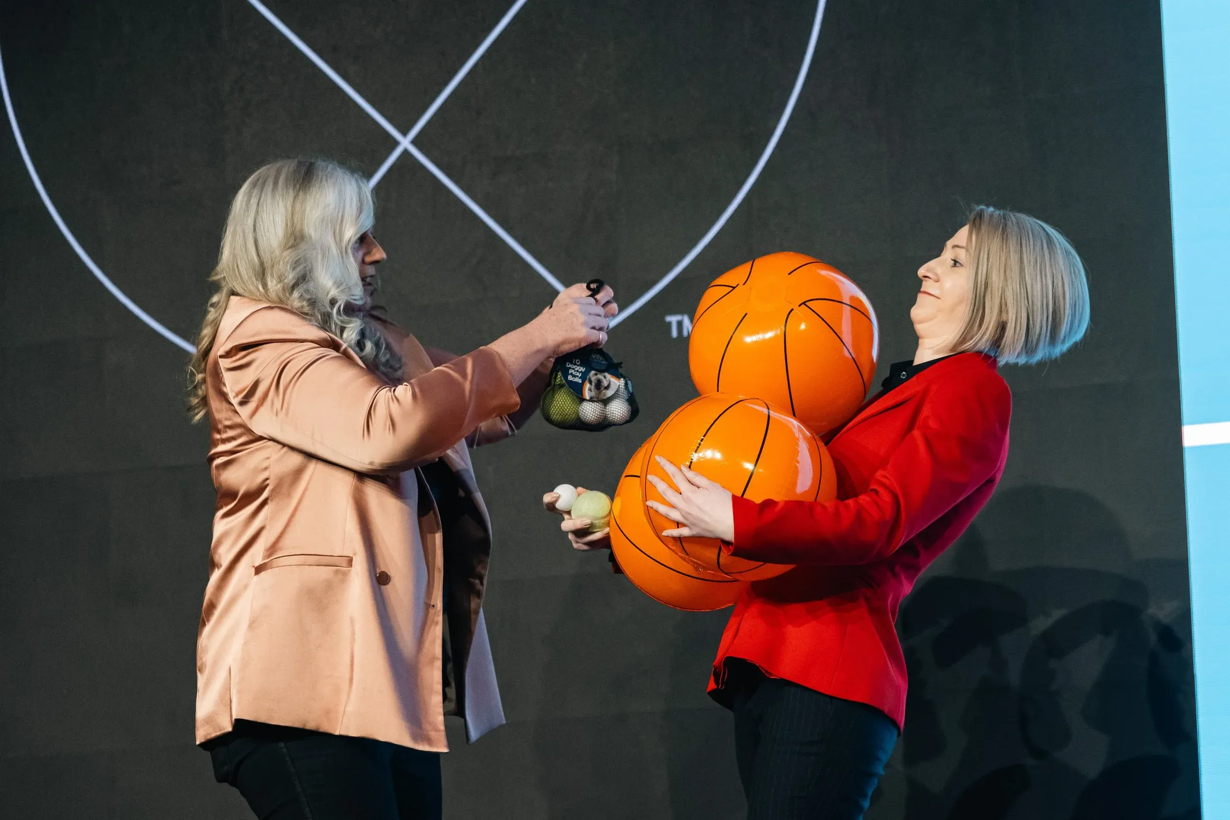 Dr Alison Edgar MBE on stage handing a colleague a net of small balls while the colleague hugs two inflatable basketballs, demonstrating the Big Balls® prioritisation exercise.