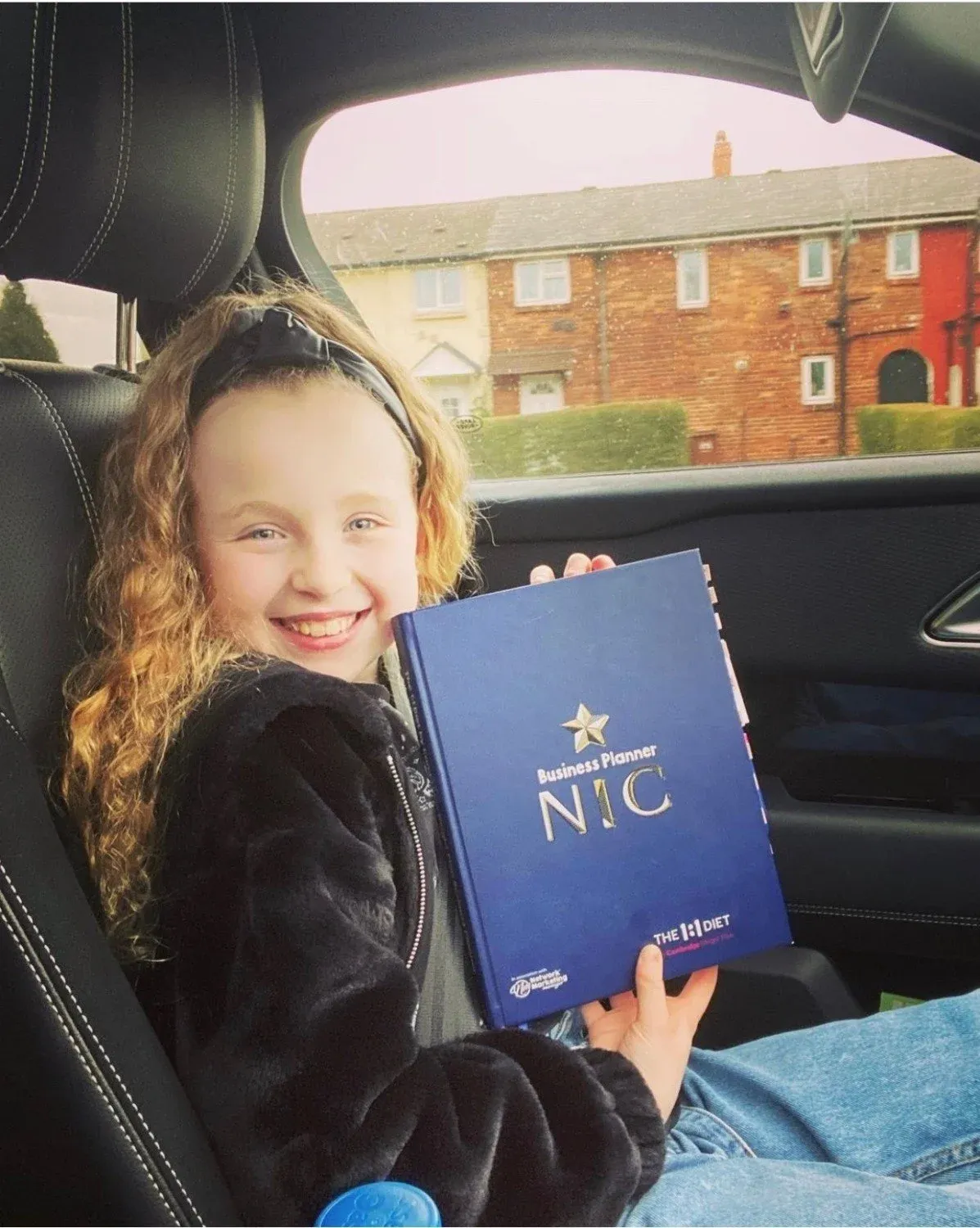 A young girl in a car seat grinning as she holds up a blue hardback business planner embossed with the name Nic.