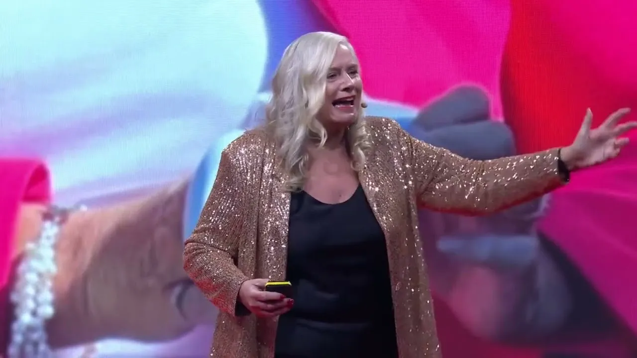 Dr Alison Edgar MBE on stage in a gold sequinned jacket, arm outstretched to the audience, at Cambridge 1:1 Diet Live.