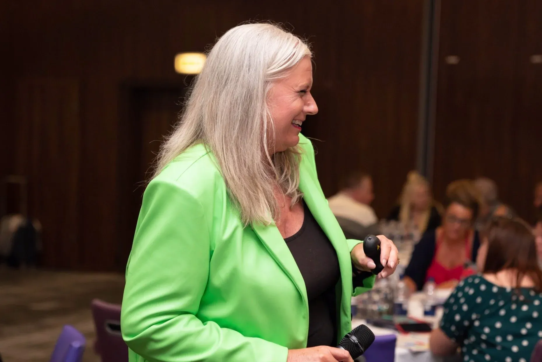 Dr Alison Edgar MBE in a bright green blazer, laughing as she works the room with a microphone in one hand and a clicker in the other.