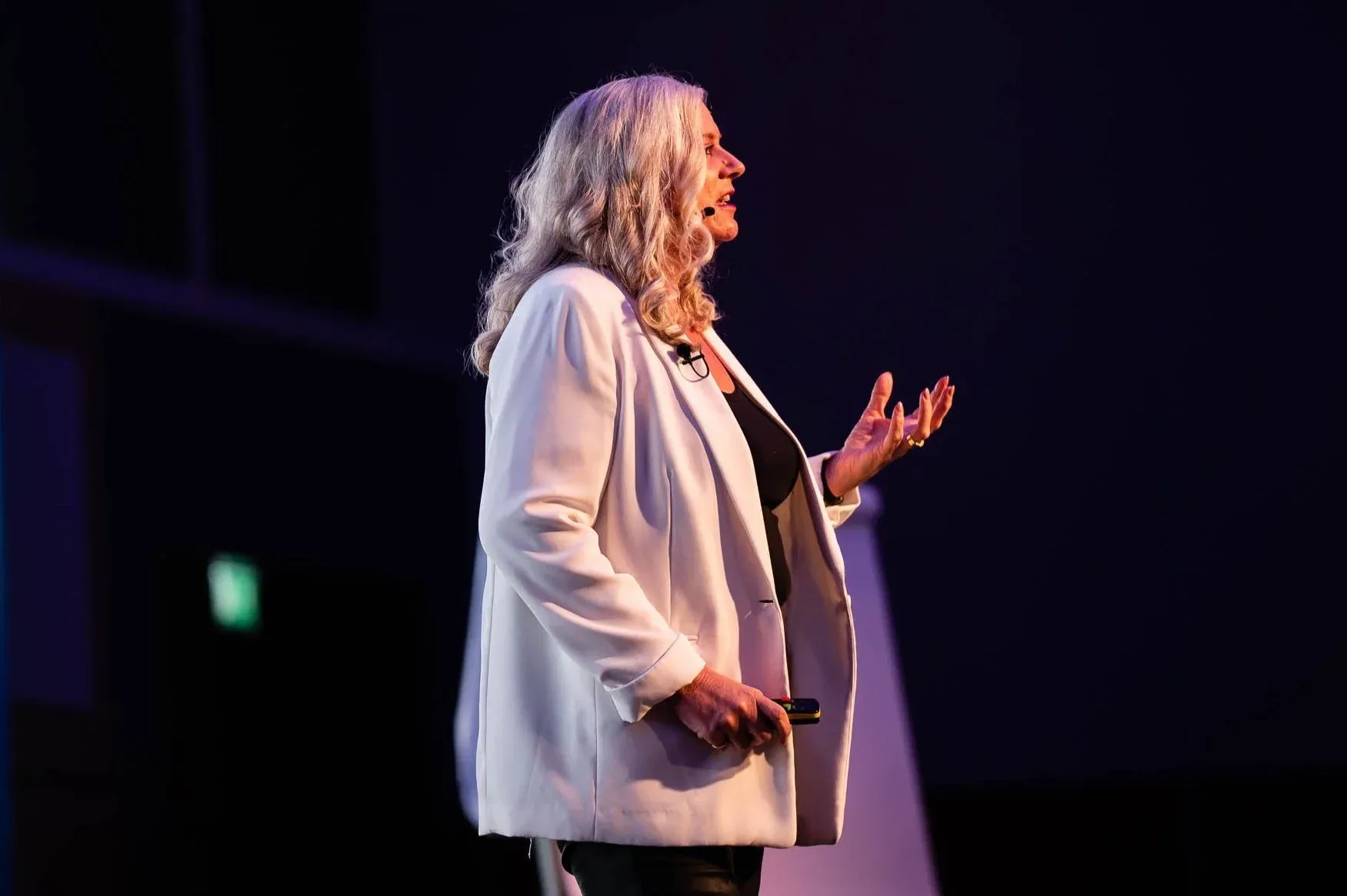 Dr Alison Edgar MBE in a white blazer on a darkened stage, one hand open as she makes a point.