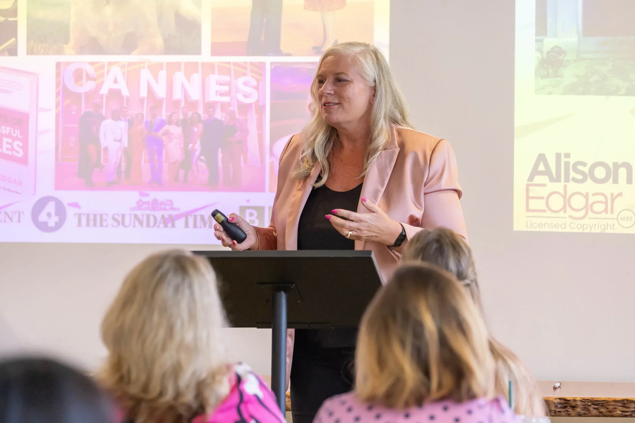 Dr Alison Edgar MBE speaking from a lectern to a seated audience, her slide behind her showing press logos and a photo from Cannes.