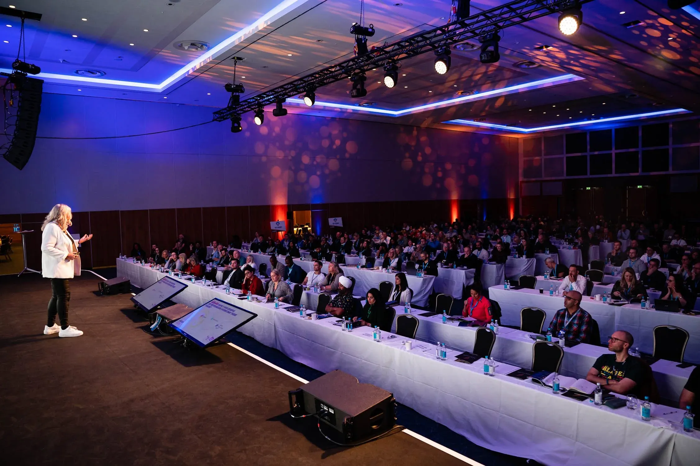 Dr Alison Edgar MBE speaking to a large conference audience seated at long tables under blue and orange stage lighting.