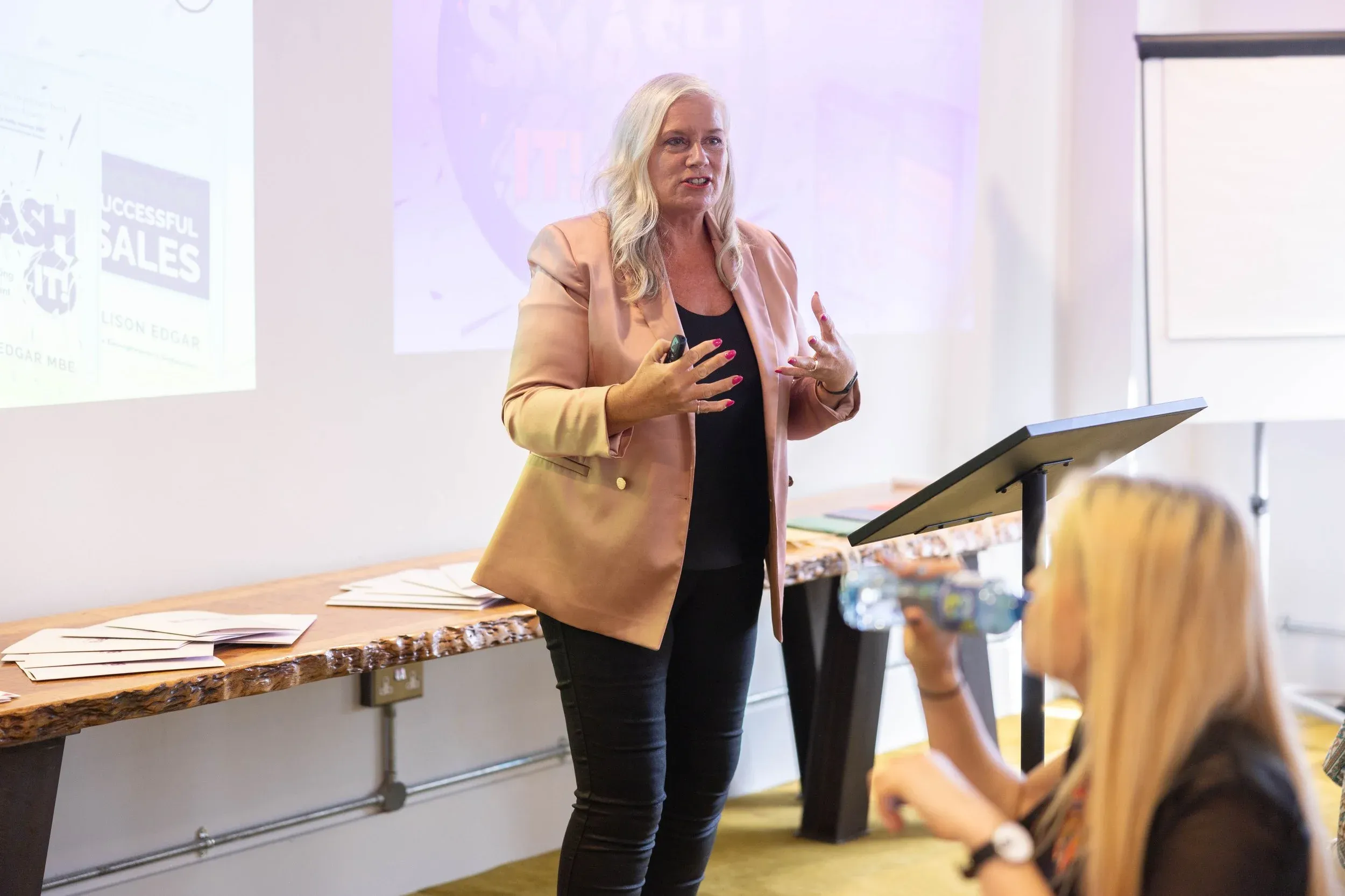 Dr Alison Edgar MBE speaking to a seated group, her book covers for SMASH IT! and Secrets of Successful Sales projected on the screen behind her.
