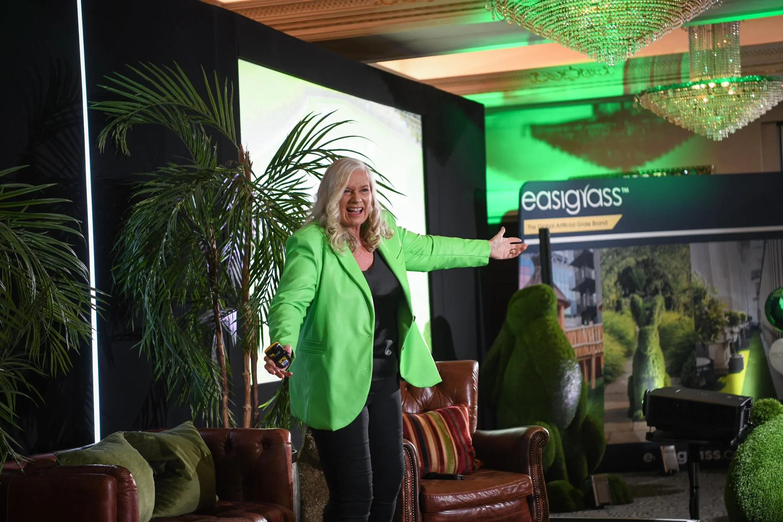 Dr Alison Edgar MBE in a green blazer, arm outstretched, presenting at an Easigrass event among palms and topiary.
