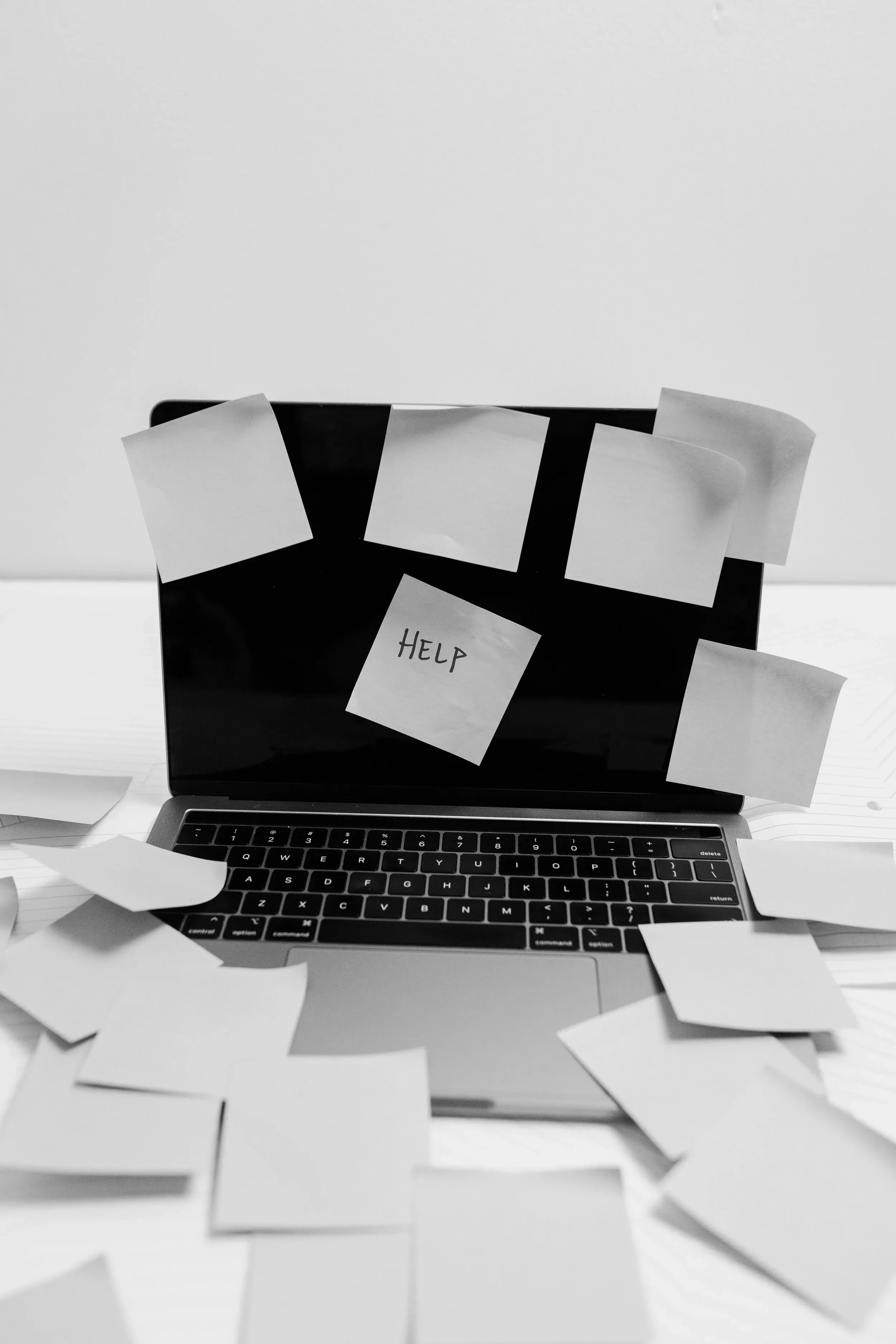 A laptop covered in blank sticky notes with one reading ‘HELP’, more notes scattered across the desk around it.